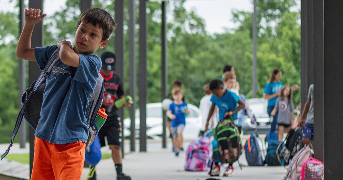 alt=Photo of a group of children carrying backpacks outside of a school. The focus is on a boy with tan skin on the far left wearing a blue shirt and orange shorts. He is in the process of putting his gray backpack on, with his right arm already through one side.